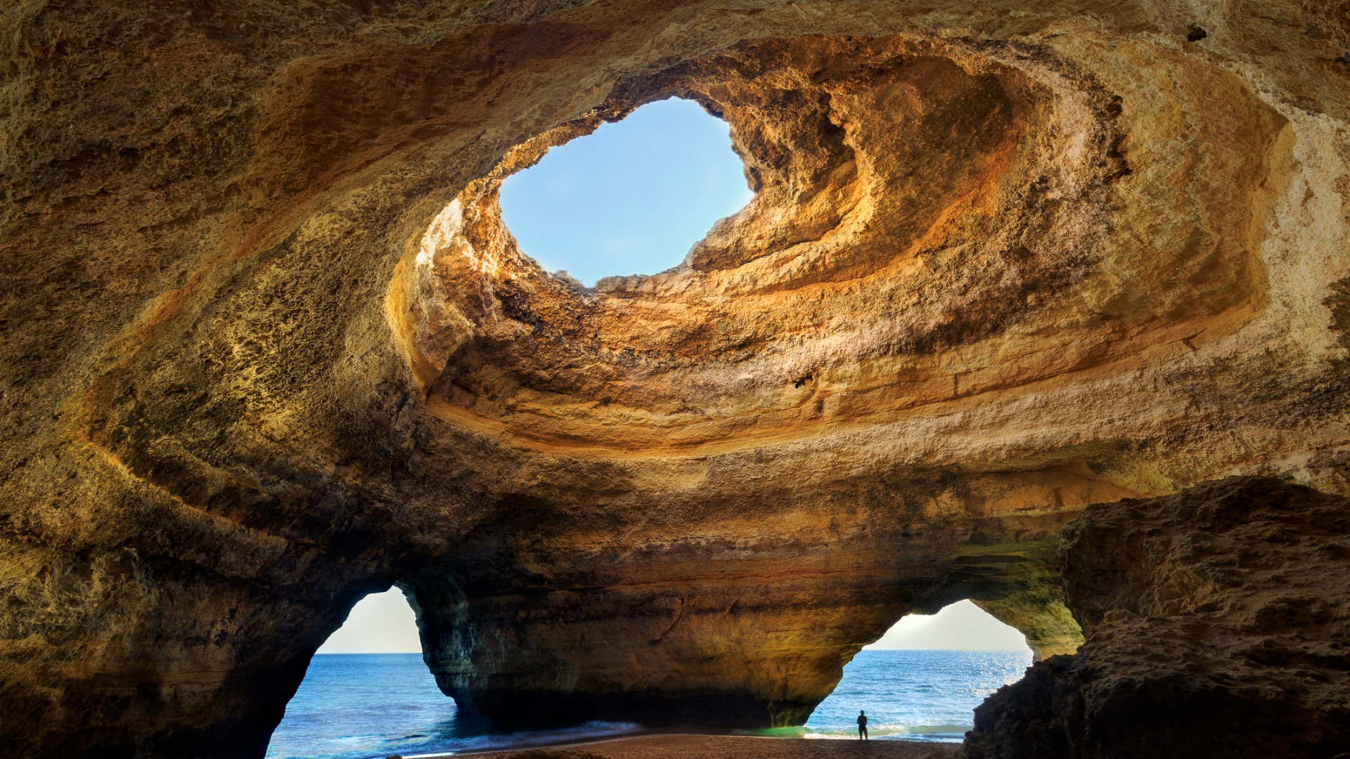 An elaborate rock formation with three large openings. A person is standing on the sand under the rock, facing the sea. 