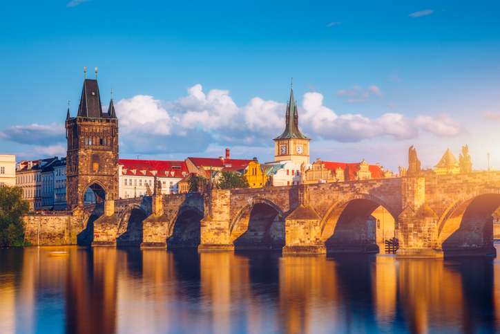 A beautiful view of the historic Charles Bridge in Prague, Czech Republic, captured during the golden hour. The bridge, with its stone arches and iconic Gothic towers, spans the calm Vltava River, reflecting the warm hues of the setting sun. The picturesque scene is complemented by the classic architecture of the surrounding buildings, bathed in soft sunlight under a clear blue sky with scattered clouds. This image highlights the timeless charm and serene beauty of one of Prague's most famous landmarks.