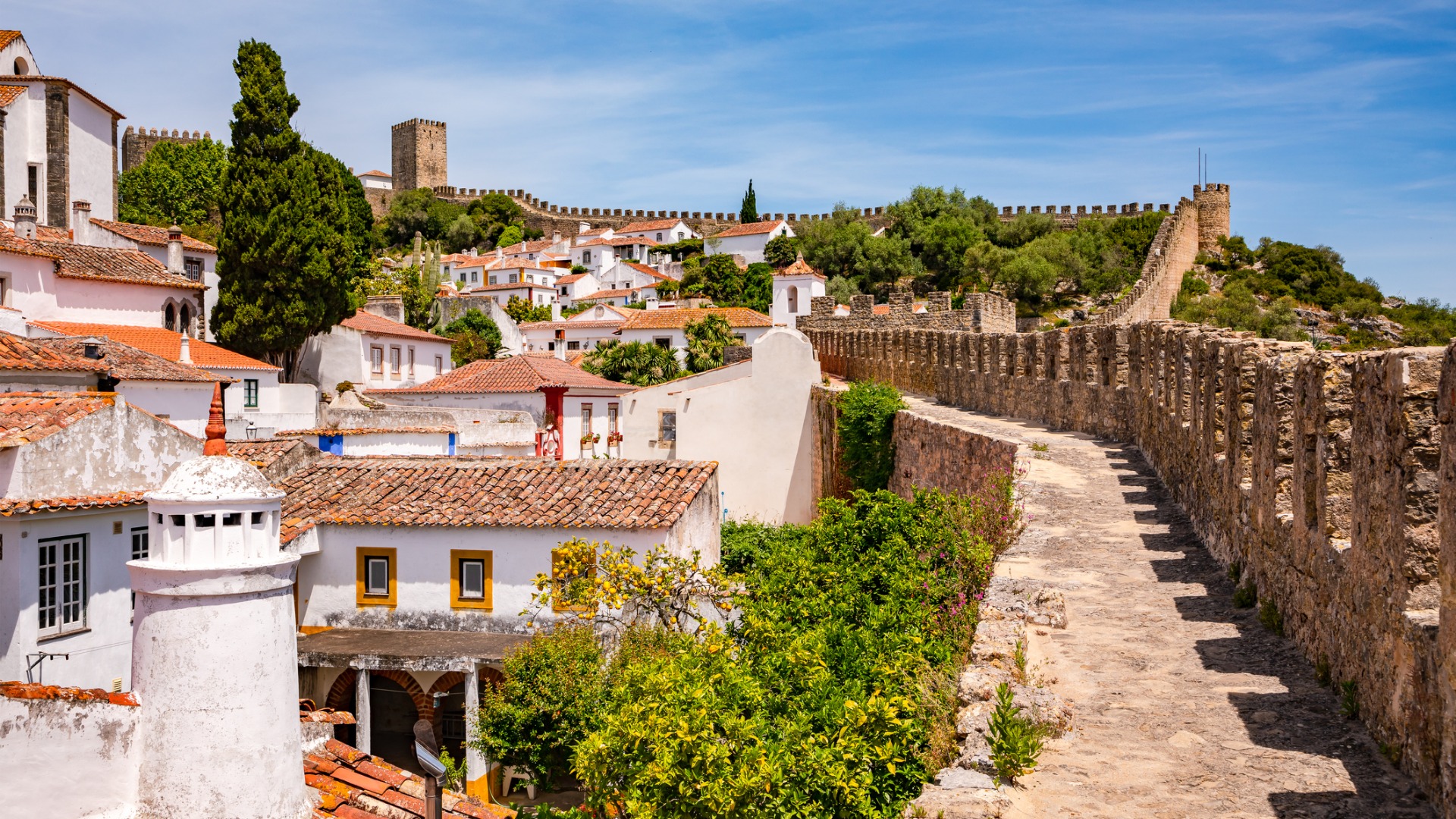 Picturesque whitewashed buildings with red-tiled roofs. On the right, the old city walls of Obidos, one of the best places to visit in Portugal. 