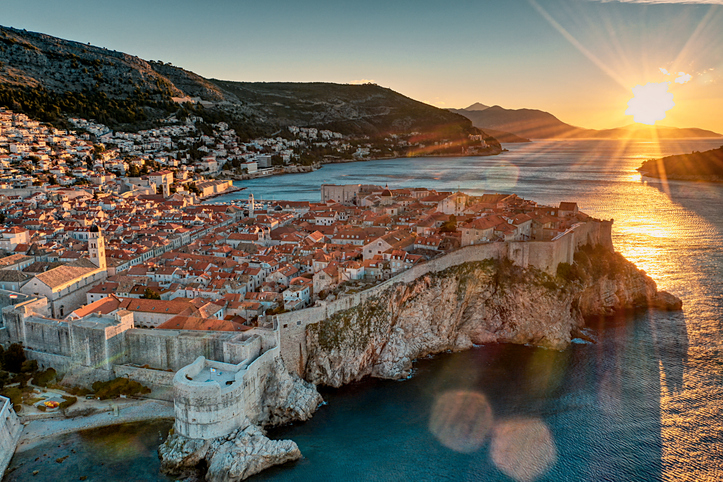 A stunning aerial view of Dubrovnik, Croatia, captured at sunset. The medieval walled city, with its distinctive terracotta rooftops, extends along a rugged coastline, surrounded by the shimmering waters of the Adriatic Sea. The golden rays of the setting sun cast a warm glow over the city, creating a picturesque scene that highlights both the natural beauty and historic charm of this UNESCO World Heritage site. The sun's reflection on the water adds a touch of brilliance to this breathtaking coastal landscape.