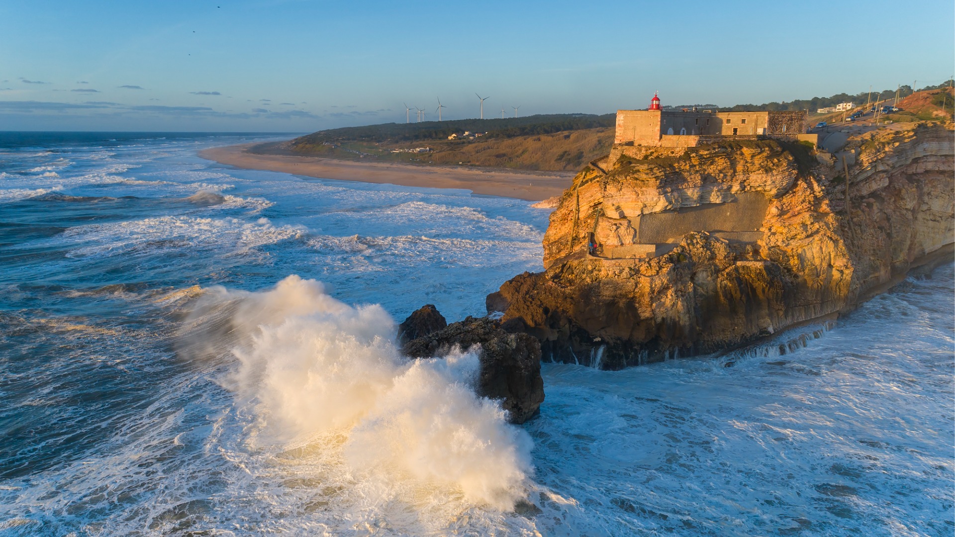 Aerial view of a lighthouse on a cliff with a fortress on the coast of the Atlantic Ocean with big waves at sunset in Nazare, one of the best places to visit in Portugal.

