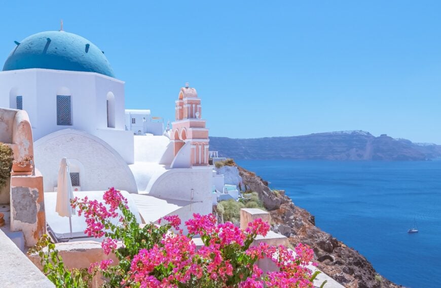 Iconic view of the blue-domed church in Oia in Santorini, with the caldera in the background and bright pink flowers in the foreground.