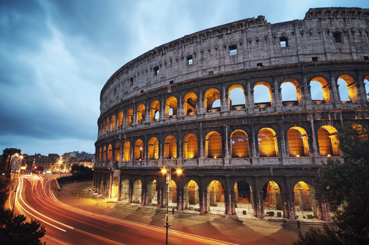 A stunning twilight view of the Colosseum in Rome, Italy, as evening sets in. The ancient amphitheater's iconic arches are illuminated with a warm golden glow, contrasting against the deepening blue of the sky. Light trails from passing vehicles create dynamic streaks around the Colosseum, adding a sense of motion to this historic setting. The scene captures the timeless grandeur of this Roman landmark, blending the old world with the energy of the modern city.