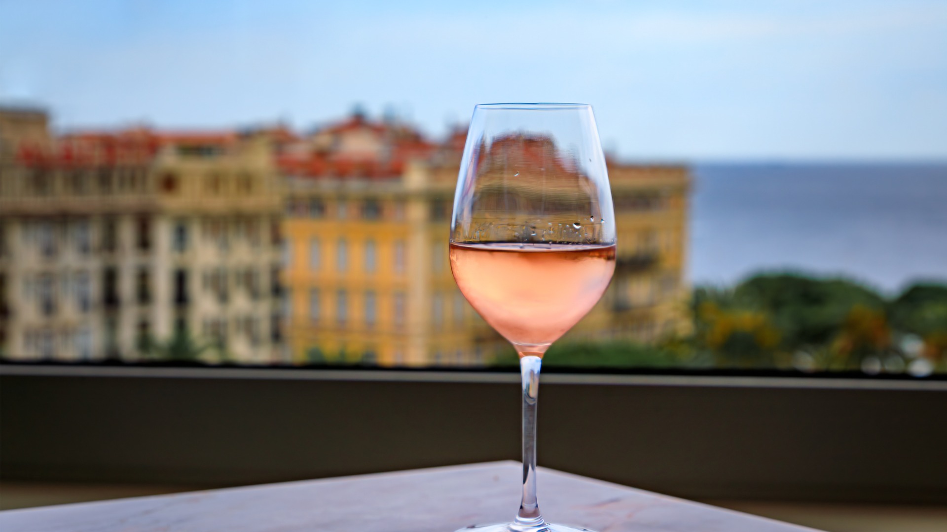 A glass of Provencal rosé wine at a rooftop bar above Nice Old Town with blurred buildings and the Mediterranean Sea in the background. 