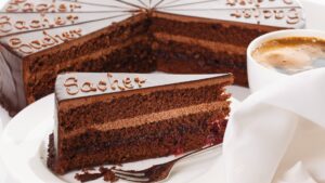 A close up of a slice of chocolate cake with the word Sacher written on it. In the background, the rest of the cake and a cup of coffee.