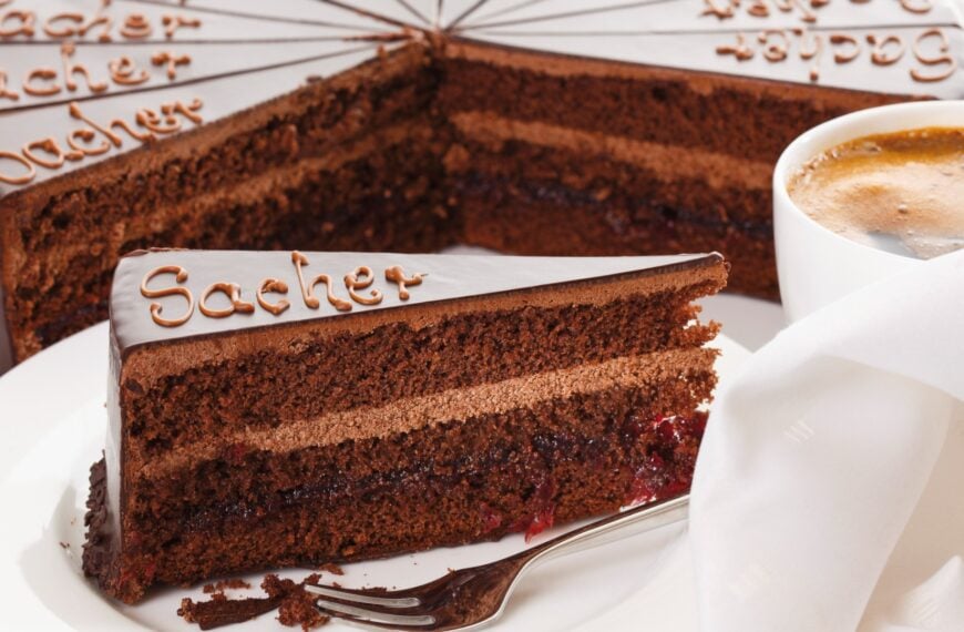 A close up of a slice of chocolate cake with the word Sacher written on it. In the background, the rest of the cake and a cup of coffee.