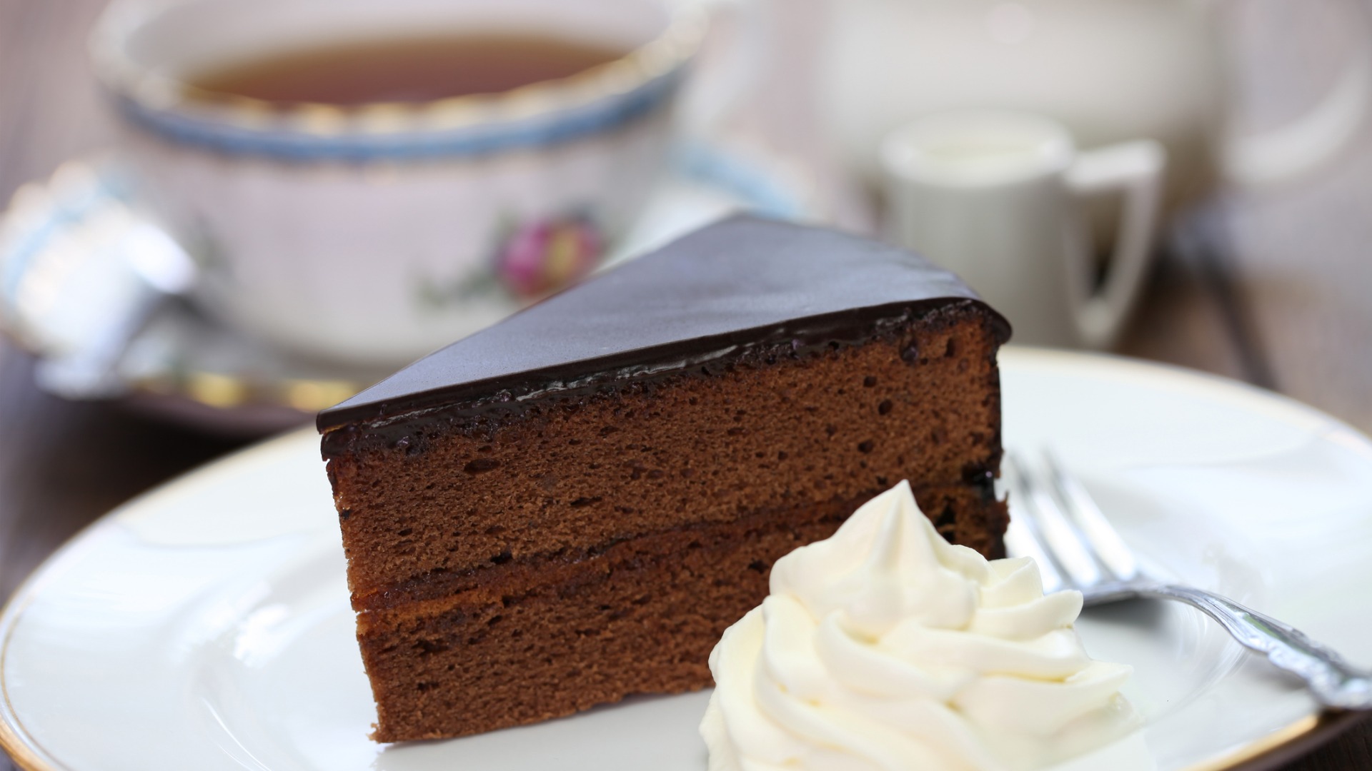 A close-up of a slice of Sachertorte with whipped cream on the side. In the background, a beautiful cup of tea. 