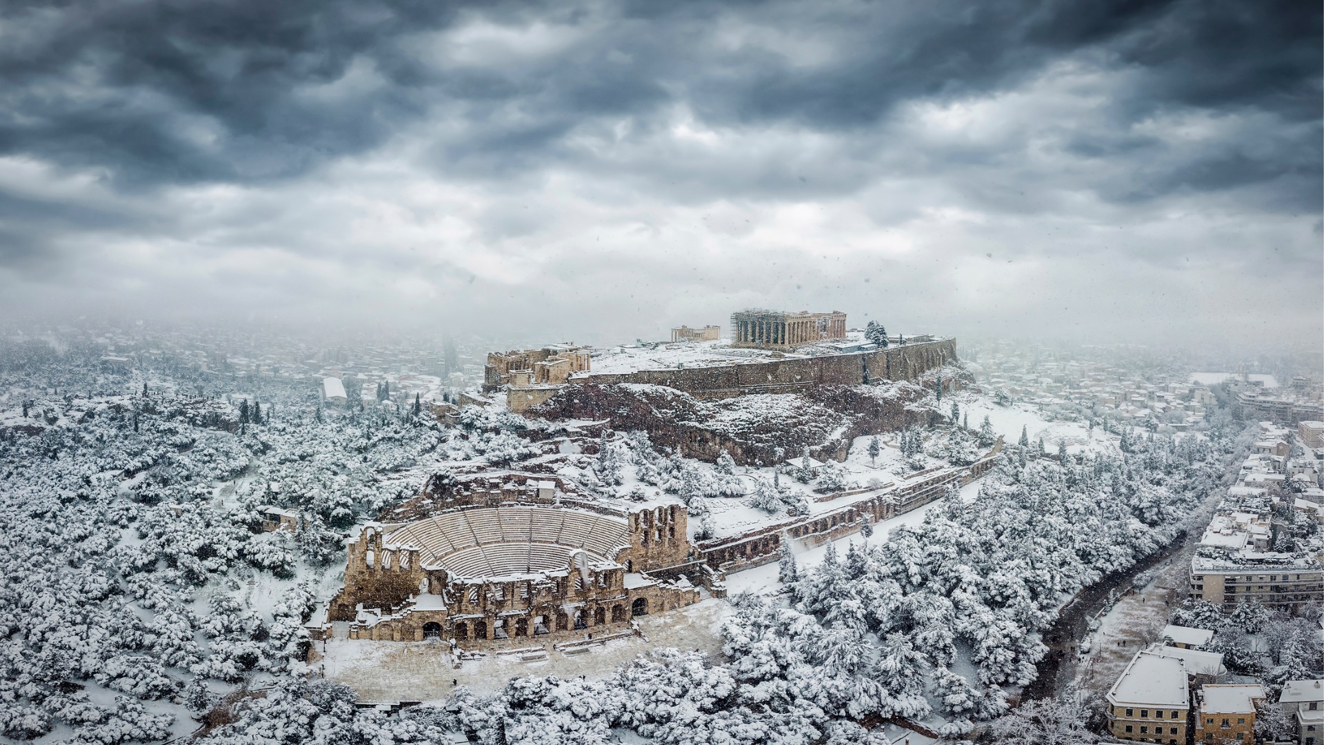 An aerial view of the Acropolis of Athens covered in snow under a dramatic sky. 