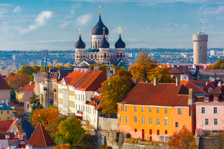 A picturesque view of Tallinn, Estonia, showcasing the colorful buildings and rooftops of the Old Town during autumn. The scene is dominated by the majestic Alexander Nevsky Cathedral with its distinctive black onion domes and golden crosses, rising above the vibrant foliage. In the background, a medieval stone tower adds to the historic charm of the city. The mix of warm autumnal colors and the rich architectural heritage create a captivating and serene landscape.