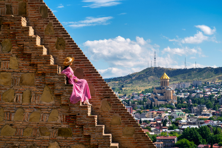 A captivating image of a woman in a flowing pink dress and straw hat sitting on the ancient stone steps of a historic fortress, overlooking the cityscape of Tbilisi, Georgia. The composition beautifully captures the contrast between the rugged, time-worn bricks of the fortress and the vibrant, modern city below. The iconic Holy Trinity Cathedral, with its golden dome, stands out in the distance against the backdrop of rolling hills and a bright blue sky dotted with fluffy clouds. This scene blends cultural heritage with the scenic beauty of Tbilisi.