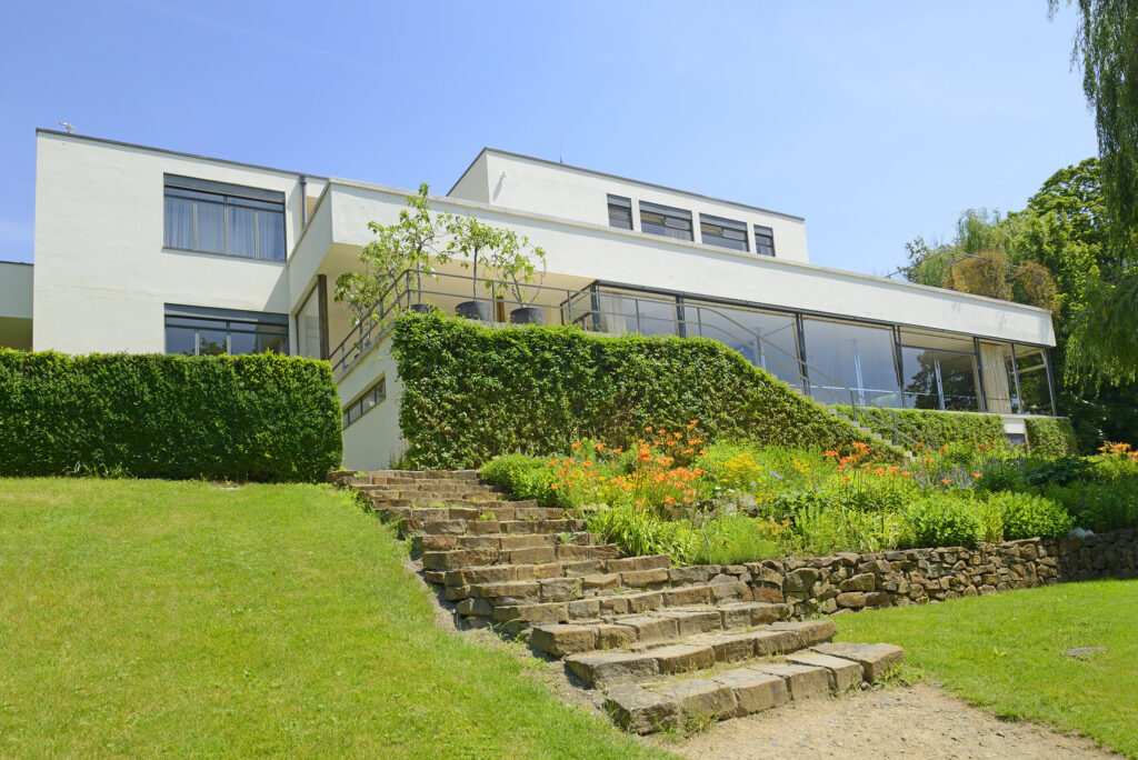 A photograph of the Villa Tugendhat, a modernist architectural masterpiece located in Brno, Czech Republic. The building features clean lines and a minimalist design with large glass windows, surrounded by well-manicured gardens. A stone staircase leads up to the house, bordered by lush greenery and colorful flowers, blending the structure seamlessly into its natural surroundings. 