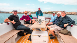 This image depicts a group of seven older adults sitting on a boat, enjoying a casual outing. They are seated around a long table, and there is a large cooler in the center. Most of them are smiling and appear relaxed, dressed in casual clothing suitable for the weather. In the background, there is a distant view of a city skyline across the water, and the sky is partly cloudy, creating a serene atmosphere for their boating trip. The overall mood is friendly and leisurely.