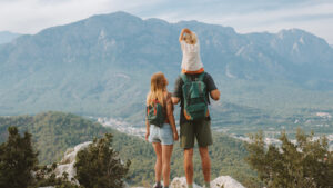 This image shows a family of three standing on a scenic mountain overlook. The man is carrying a child on his shoulders, while a woman stands beside him. All three are wearing backpacks, indicating they are likely on a hike. They are facing away from the camera, gazing at the vast mountain range in the distance, with greenery and a small town visible below. The scene exudes a sense of adventure, exploration, and togetherness, with the family enjoying a peaceful moment in nature.