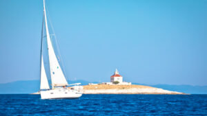 This image captures a serene scene of a white sailboat gliding across the deep blue water. In the background, there is a small island with a quaint lighthouse standing on its rocky shore. The sky is clear and bright, complementing the calmness of the sea. The image evokes a peaceful, idyllic atmosphere, with the sailboat and lighthouse creating a picturesque maritime setting.