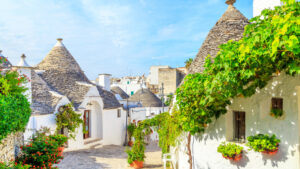 This image depicts a charming village with traditional whitewashed buildings topped with unique conical roofs, known as trulli. The narrow cobblestone street is lined with vibrant potted plants and greenery, giving the scene a bright and inviting feel. Sunlight bathes the village, highlighting the quaint architecture and lush vines draping over some of the houses. The overall atmosphere is peaceful and picturesque, evoking the charm of a historical Mediterranean town.