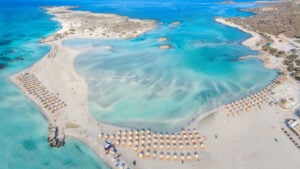 This image shows a stunning aerial view of a beach with crystal-clear turquoise waters and soft sandy shores. Rows of sun umbrellas are neatly arranged along the beach, offering a picturesque, organized setting for beachgoers. The shallow water extends in beautiful shades of blue, creating a peaceful lagoon surrounded by small rocky islets. The scene is framed by the natural beauty of the coastline, with a mix of sandy stretches and rocky outcrops. The tranquil and inviting atmosphere makes this beach a perfect destination for relaxation and leisure.