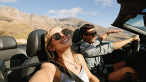 This image shows a young couple enjoying a road trip in a convertible. The woman in the passenger seat is taking a selfie, smiling brightly while wearing sunglasses, and the man driving is laughing, pointing toward something in the distance. They are driving through a scenic landscape, with rugged mountains and a clear blue sky visible in the background. The open roof and sunshine create a sense of freedom, adventure, and joy as the couple enjoys their journey together.