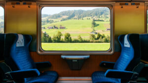 This image shows the view from the interior of a train, looking out through a large window. Inside, two comfortable blue seats face each other, creating a cozy and inviting setting. Through the window, a beautiful countryside scene unfolds, with rolling green hills, scattered trees, and a few quaint houses in the distance. The sunlight enhances the vibrant colors of the landscape, creating a peaceful and scenic travel moment, perfect for a leisurely train ride through nature.