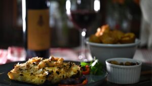 A close-up of a dish of roasted cod with a bottle of wine and a glass of red wine in the background.