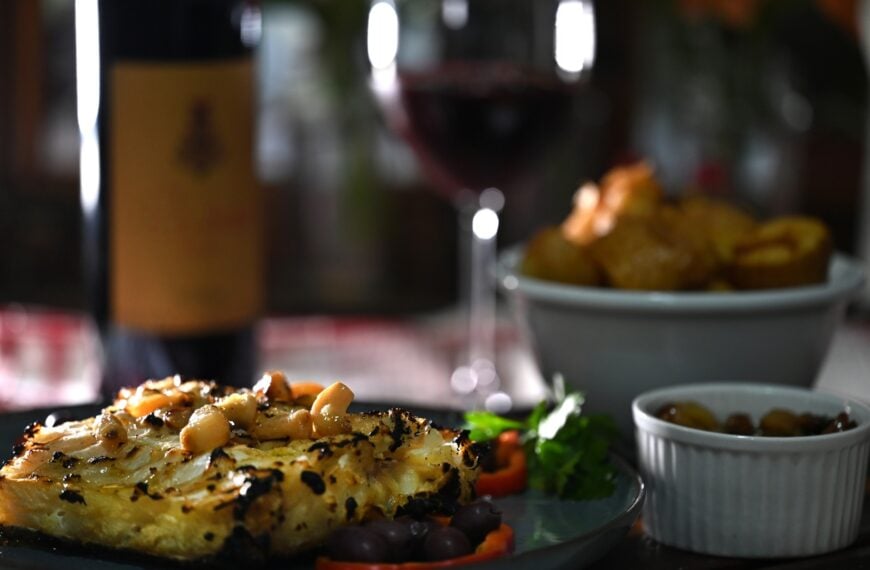 A close-up of a dish of roasted cod with a bottle of wine and a glass of red wine in the background.