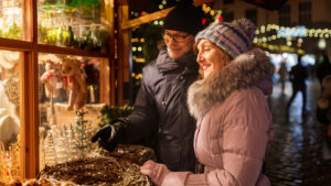 An older couple is enjoying a festive outdoor market, dressed warmly in winter coats, hats, and gloves. The woman is wearing a pink coat with a fur-lined hood and a knitted hat, while the man is in a dark coat with a black hat and scarf. They are smiling and admiring various holiday decorations displayed in a wooden stall, including small Christmas trees and ornaments, with a backdrop of warm, glowing lights that enhance the cozy, cheerful atmosphere.