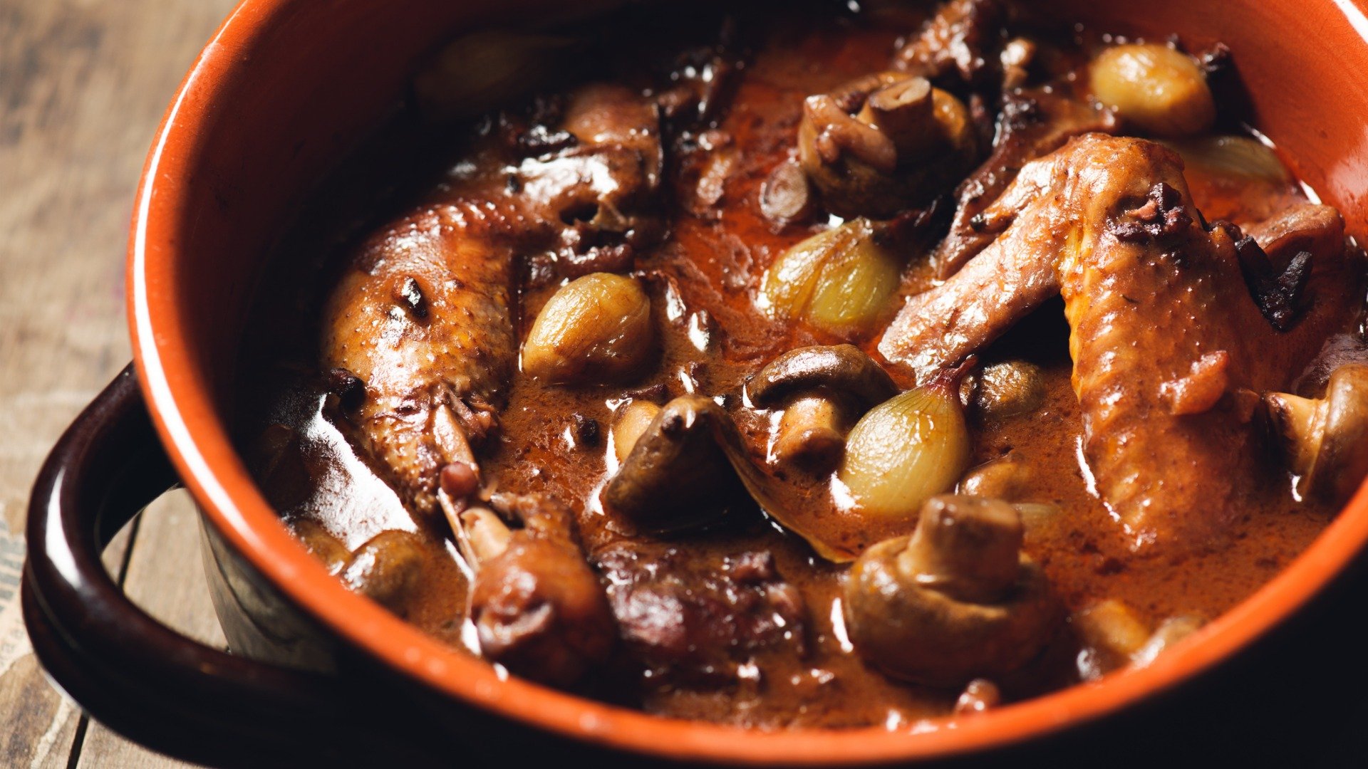 A close up of a clay pot with Coq au Vin or chicken stew with mushrooms and onions in a tomato sauce.  