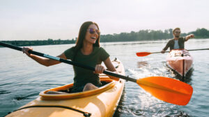 A joyful moment of two people kayaking on a calm lake. The woman in the foreground is smiling broadly as she paddles her yellow kayak, while a man in the background follows in a pink kayak. Both are enjoying the serene outdoor setting, with the water reflecting the clear sky and distant shoreline, capturing the fun and excitement of water-based adventure.