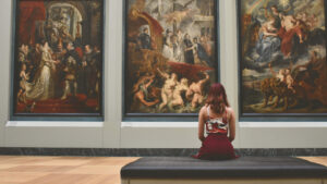 A woman sits on a bench in an art museum, gazing intently at three large, classical paintings displayed on the wall in front of her. The paintings depict historical and mythological scenes, rich with detail and vibrant colors. The woman, dressed in a stylish outfit with a patterned top and red skirt, is positioned centrally, adding a sense of contemplation and admiration for the art. The scene captures the quiet, reflective atmosphere of an art gallery, where one can immerse themselves in the beauty and history of the works on display.