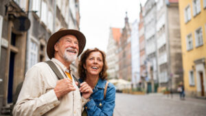 A cheerful older couple enjoying a walk through a charming European city. The man, wearing a hat and light-colored shirt with a backpack, smiles broadly while looking up, possibly admiring the architecture. The woman, dressed in a denim jacket, walks beside him, also smiling and engaged in the experience. The background features picturesque, colorful buildings lining the cobblestone street, capturing the joy of travel and exploration together.