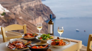 A picturesque dining scene overlooking a stunning coastal cliffside, likely in Santorini, Greece. The table is set with a delicious spread of Mediterranean cuisine, including grilled seafood, pasta with tomato sauce and seafood, a fresh salad, and a bottle of white wine with two glasses. The serene blue waters of the sea are visible in the background, adding to the ambiance of a perfect meal enjoyed with a breathtaking view.