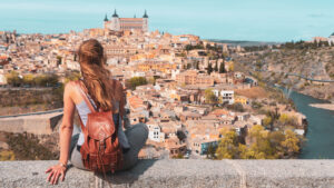 A traveler with a backpack sits on a ledge, gazing over the historic cityscape of Toledo, Spain. The view captures the city's terracotta rooftops, winding streets, and the impressive Alcázar fortress perched on a hill, all set against the backdrop of the Tagus River. The scene evokes a sense of adventure and reflection, highlighting the beauty of exploring ancient cities.