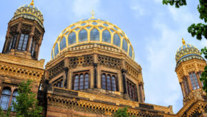 A close-up view of the New Synagogue in Berlin, Germany, featuring its stunning golden dome and intricate Moorish revival architecture. The richly detailed structure, with its ornate designs and vibrant colors, stands out against a clear blue sky.