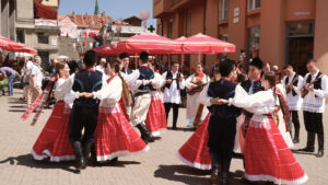 A lively street scene featuring a group of dancers in traditional Eastern European costumes performing a folk dance. The women are dressed in red and white skirts with embroidered blouses, while the men wear dark pants and vests with matching traditional attire. They are dancing in pairs, holding hands in a circle, creating a vibrant and festive atmosphere. The performance is taking place in an outdoor setting, surrounded by onlookers and cafes with red umbrellas, celebrating cultural heritage and community spirit.