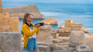 A young woman, dressed in a yellow cardigan and denim shorts, stands among ancient ruins with a camera in hand, capturing the scenic coastal views. She is focused on her photography, with the deep blue sea and rocky cliffs in the background, likely in a historic Mediterranean location. The scene reflects a blend of exploration, history, and the beauty of the natural landscape.