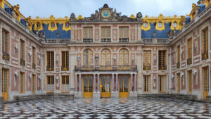 A detailed view of the ornate façade of the Palace of Versailles in France, showcasing its grand Baroque architecture. The building features elaborate decorations, including gold accents, sculpted reliefs, and large windows with yellow frames. The foreground highlights the striking black and white geometric patterned courtyard, which reflects the opulence and meticulous design of this historic royal residence. The scene captures the splendor and elegance of one of Europe's most famous palaces.