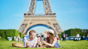 A couple enjoying a romantic picnic on the grassy lawns near the Eiffel Tower in Paris, France. They are lying on a blanket, sharing a meal that includes croissants and champagne, with a picnic basket nearby. The woman is wearing a sunhat, and both are smiling and engaged in conversation, with the iconic Eiffel Tower prominently towering in the background. The scene captures the quintessential Parisian experience, filled with love, relaxation, and the beauty of one of the world's most famous landmarks.