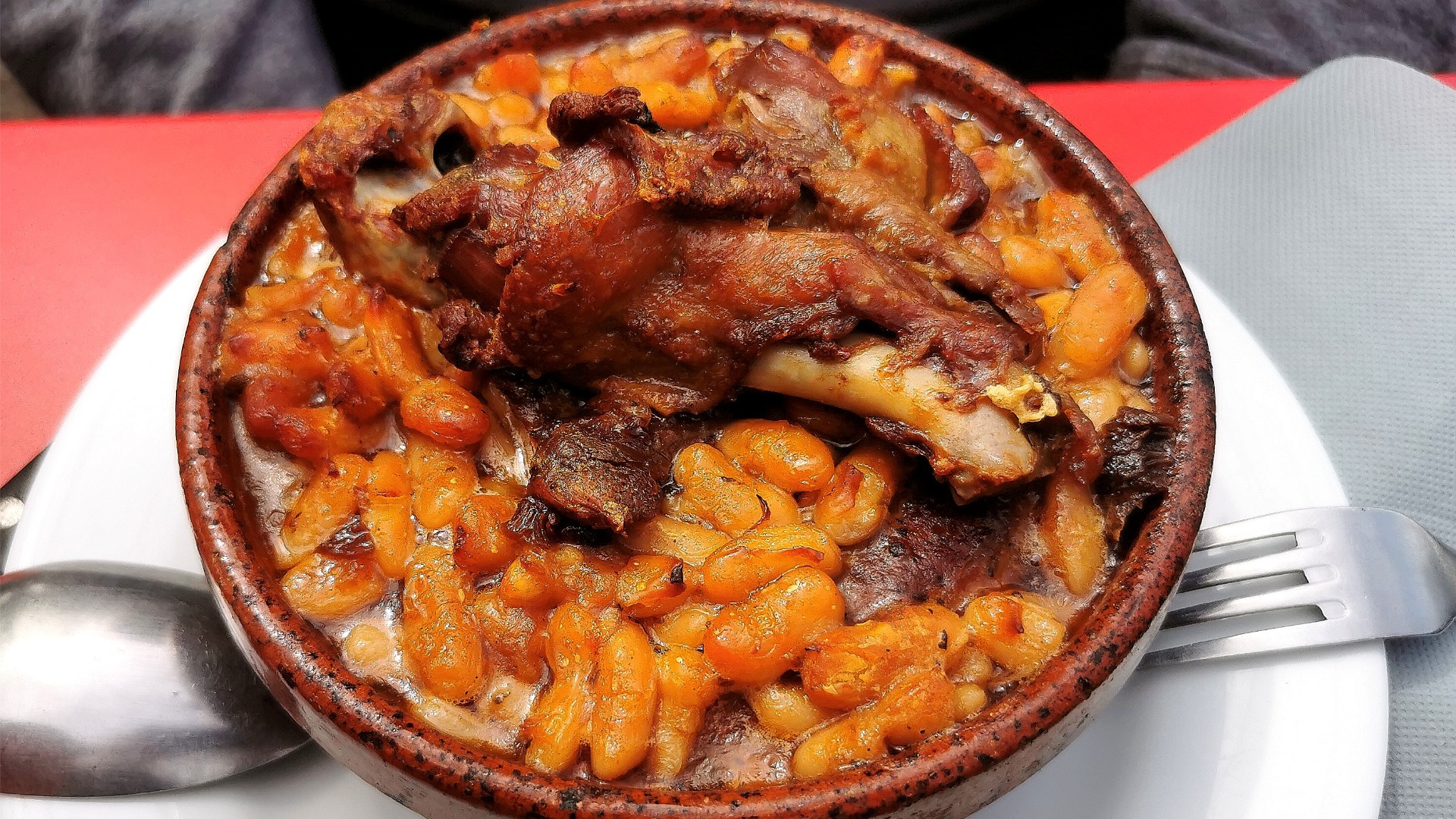 A close up of a clay dish filled with white beans in a tomato sauce and a piece of meat on top.