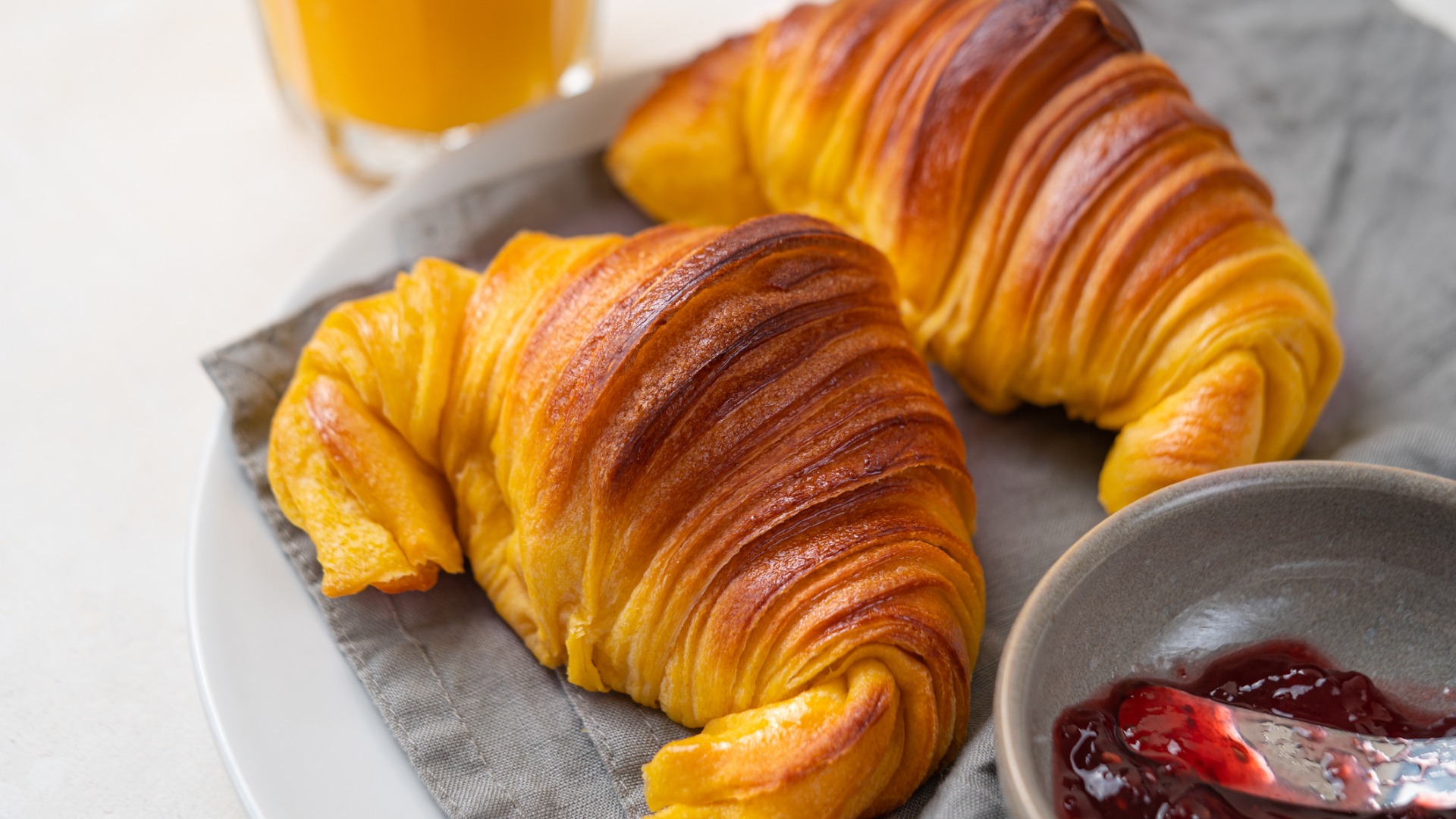 Brioche croissants on a plate with a small bowl of jam in the foreground and a glass of orange juice in the background. 