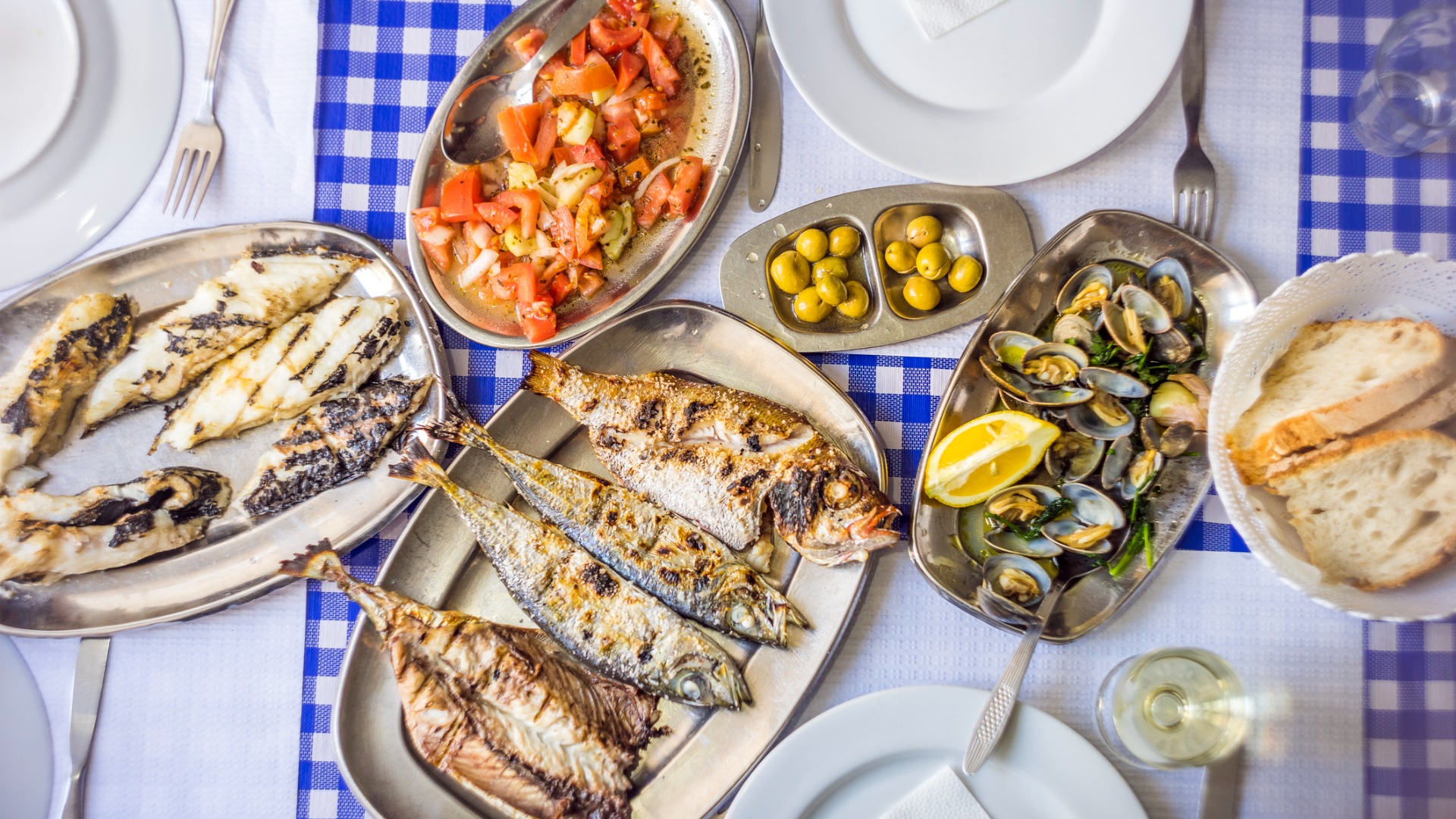 A top-down image of a table with a white and blue checkered tablecloth and several dishes of fish and seafood on top. 