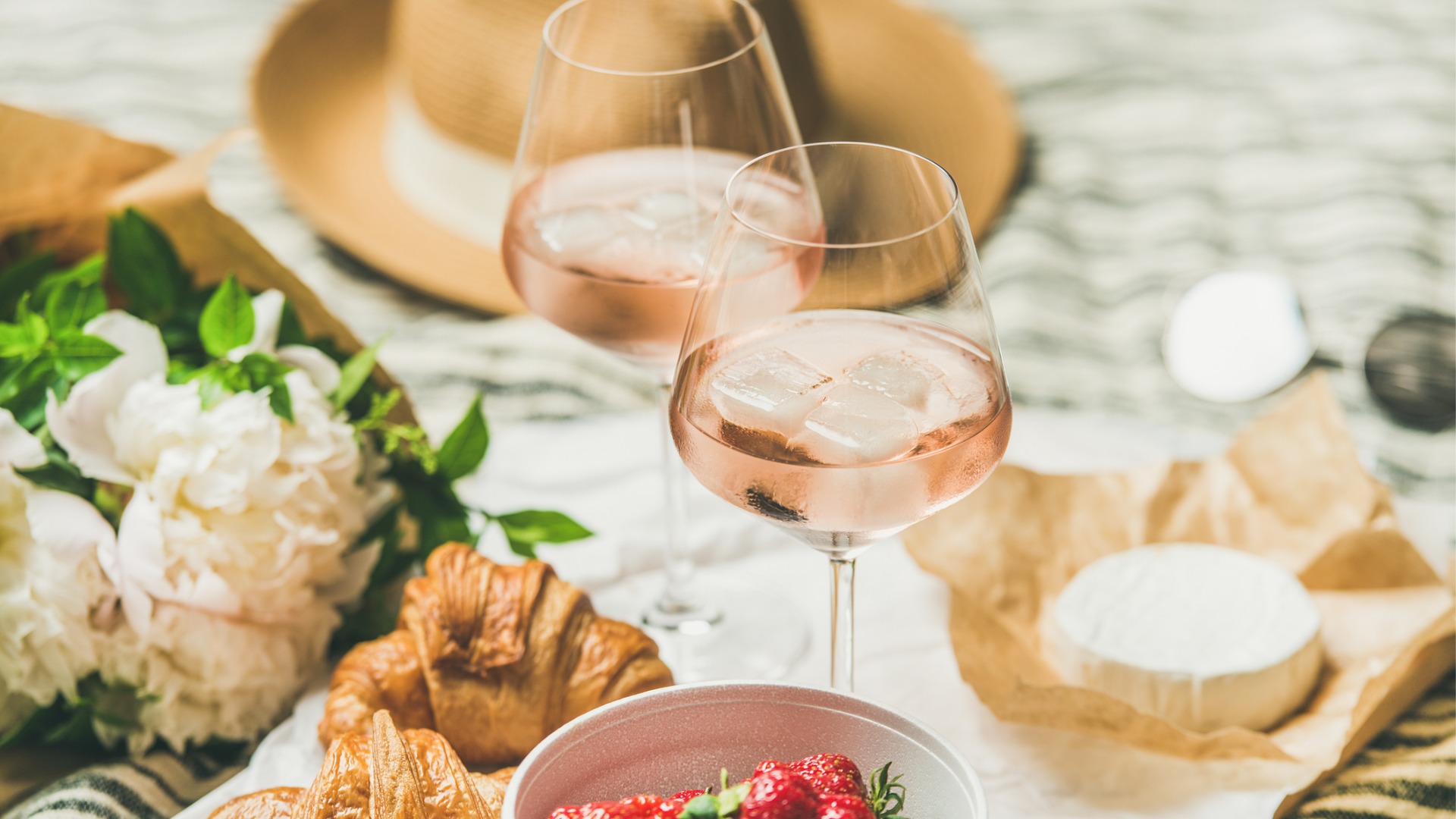 This image shows two glasses of Provencal rose wine, brie cheese, two croissants, a bowl with strawberries, white flowers, and a hat in the background. 