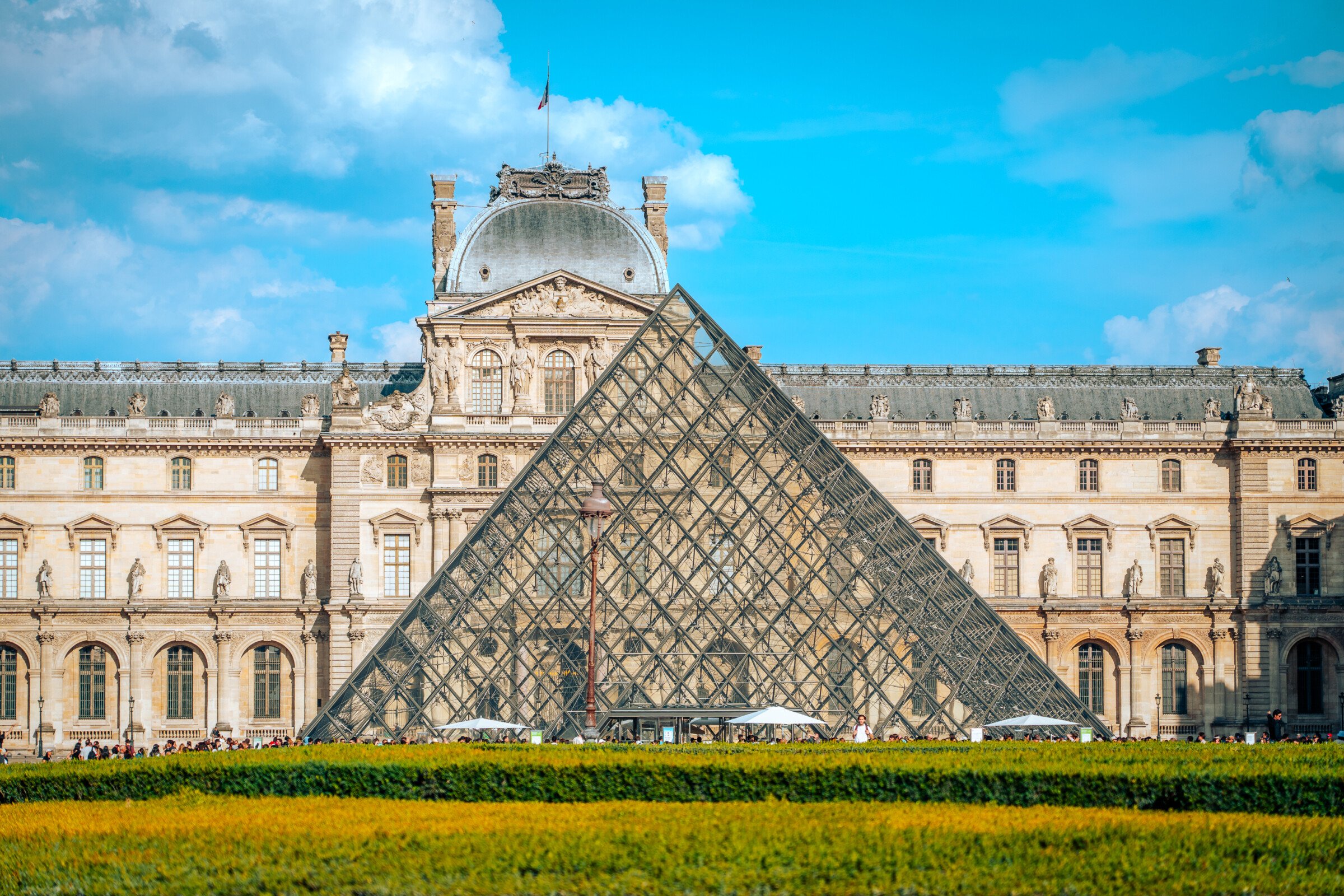 The iconic pyramid of the Louvre with the museum building in the background under a clear blue sky. 