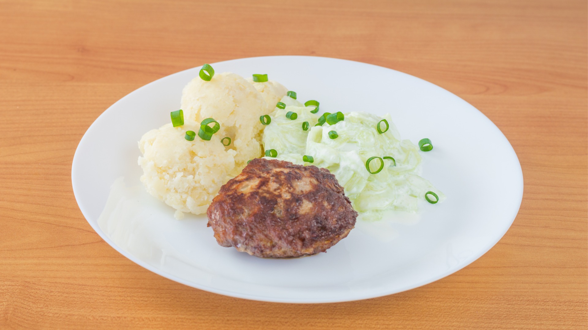 A white plate with a meat patty, mashed potatoes, and a salad made of sliced cucumbers. 