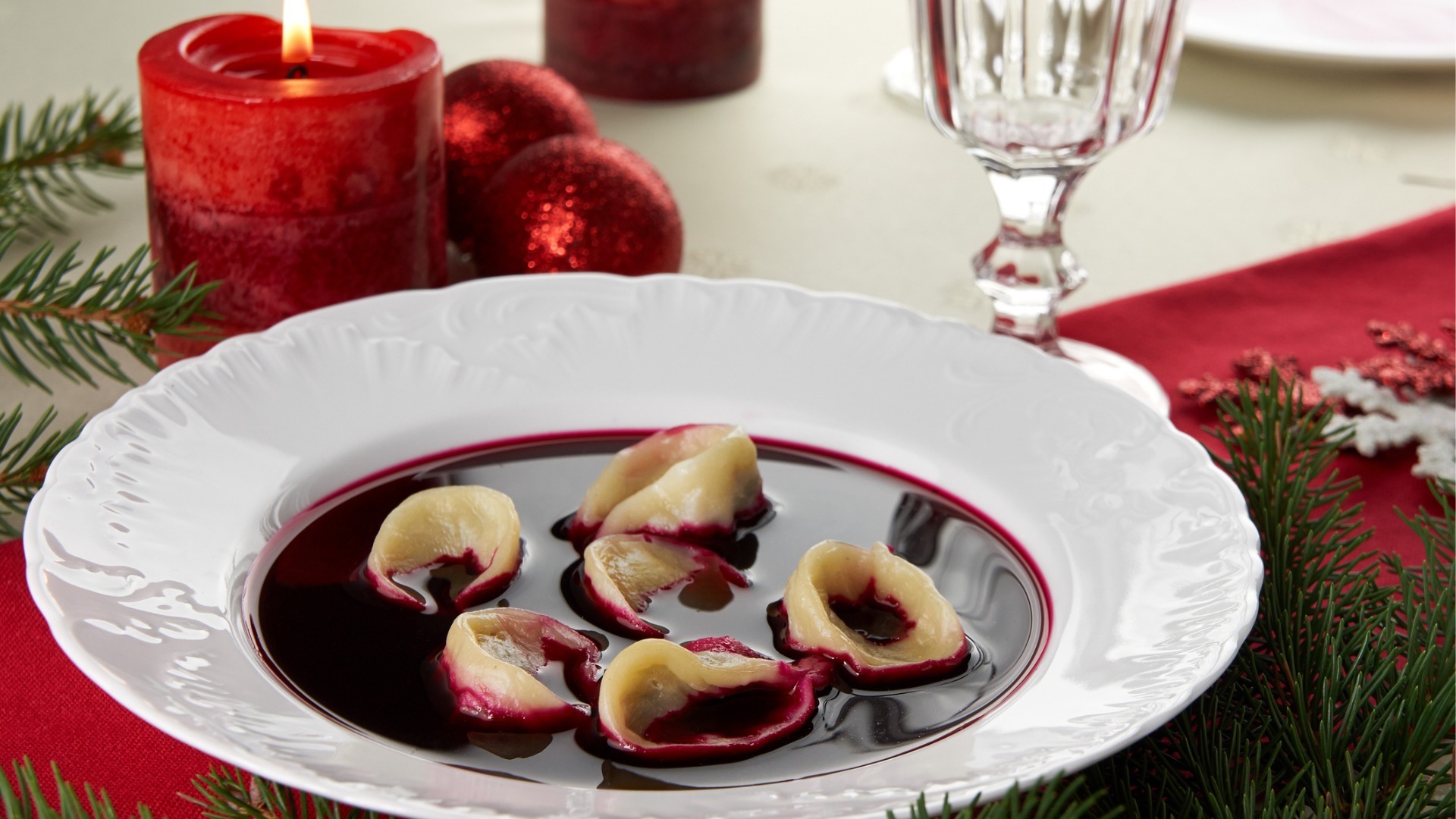 A close-up of a white plate with a red-colored soup with dumplings. A red candle in the background. 