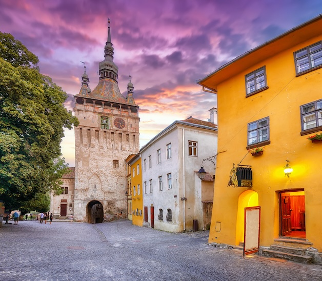 Sighisoara town square in Transylvania Romania at dusk, with a medieval clock tower, purple clouds and brightly coloured buildings.