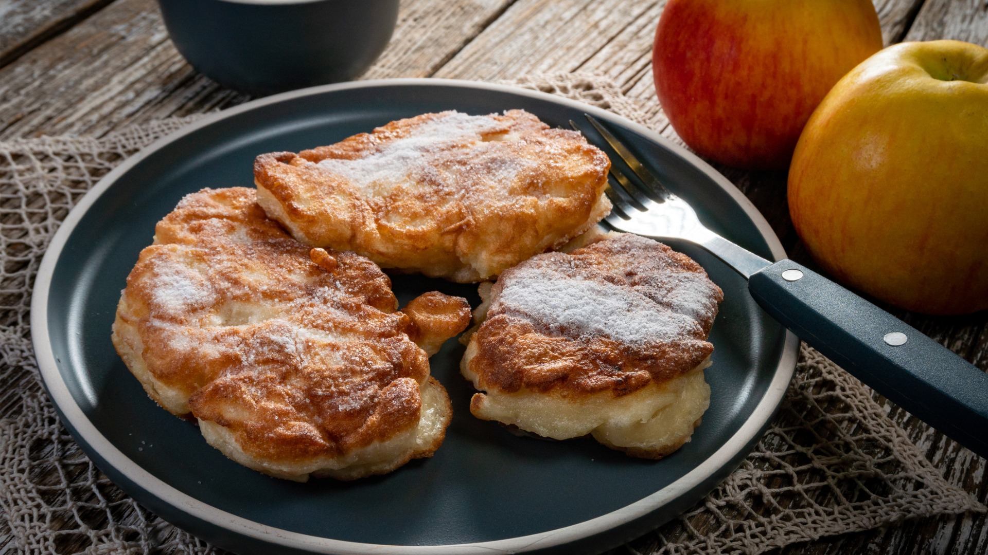 A grey plate with three large pancakes. There are two red apples next to the plate. 