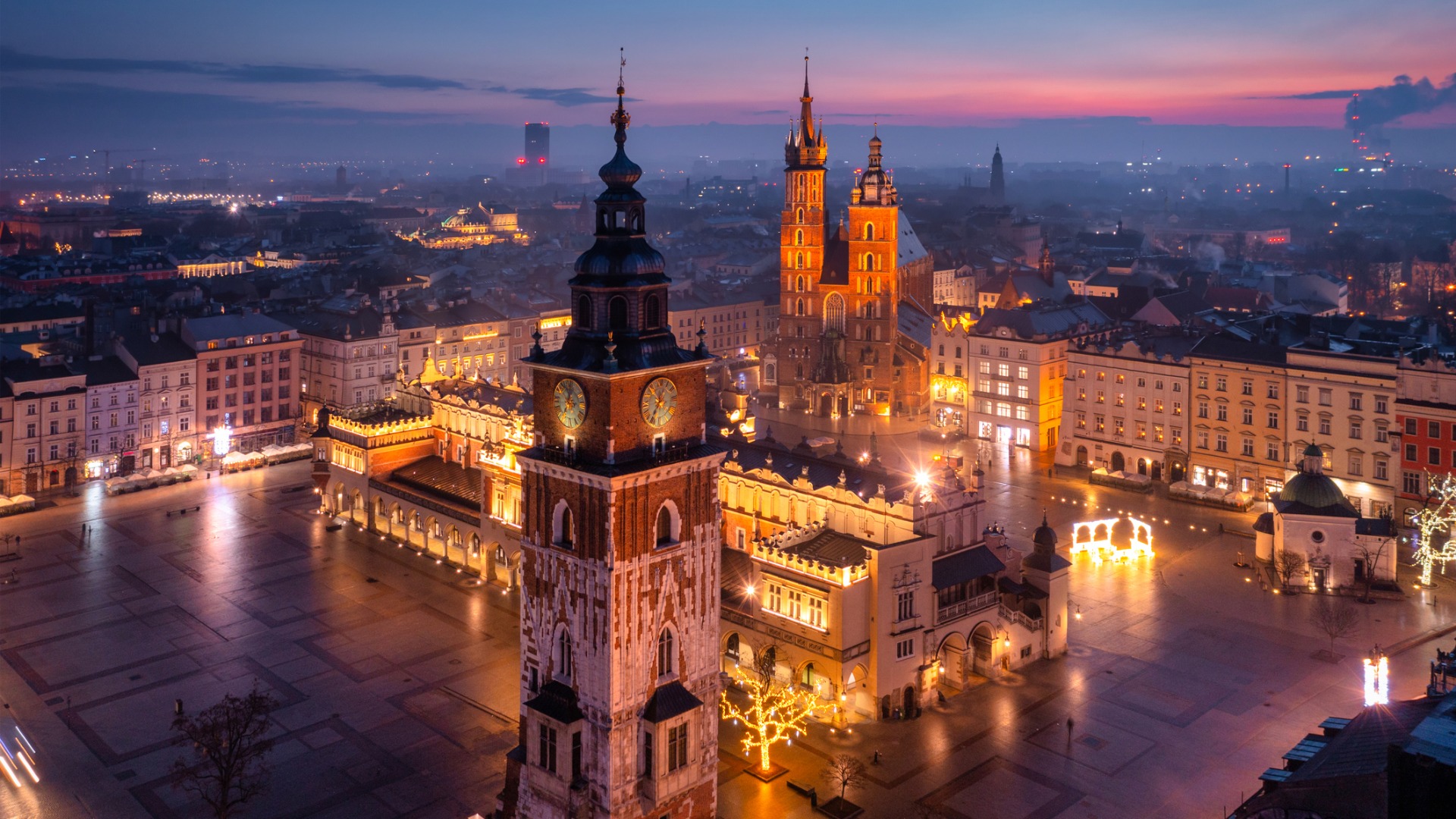 An aerial view of Krakow at sunrise. The buildings of the main square are illuminated and the sky is pink in the background. 