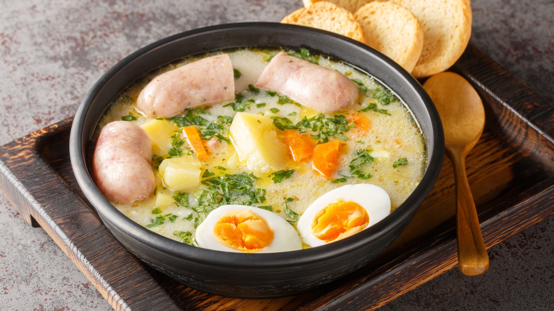 A close up of a soup with white sausage, potatoes, carrots and hard-boiled egg halves. 