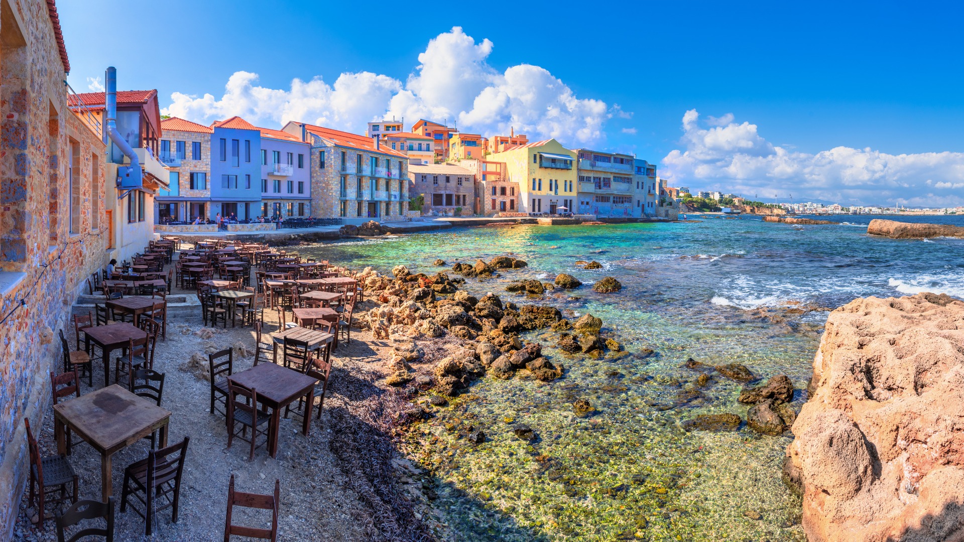 Empty chairs and tables by the sea with a row of traditional buildings in the background. 