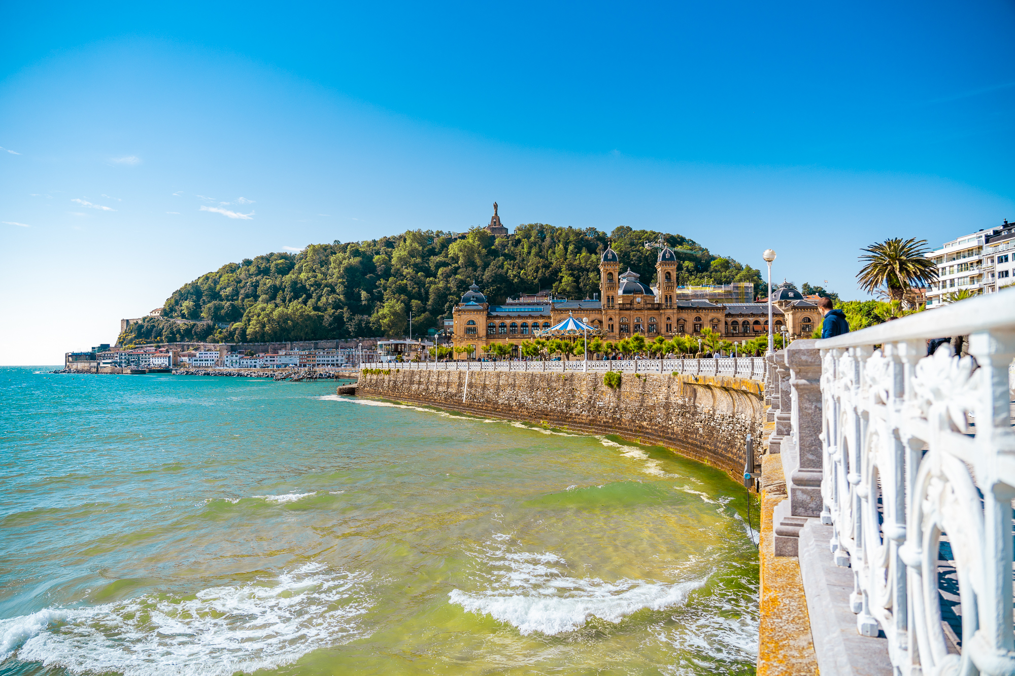 A promenade by the sea with a lush hill in the background. 