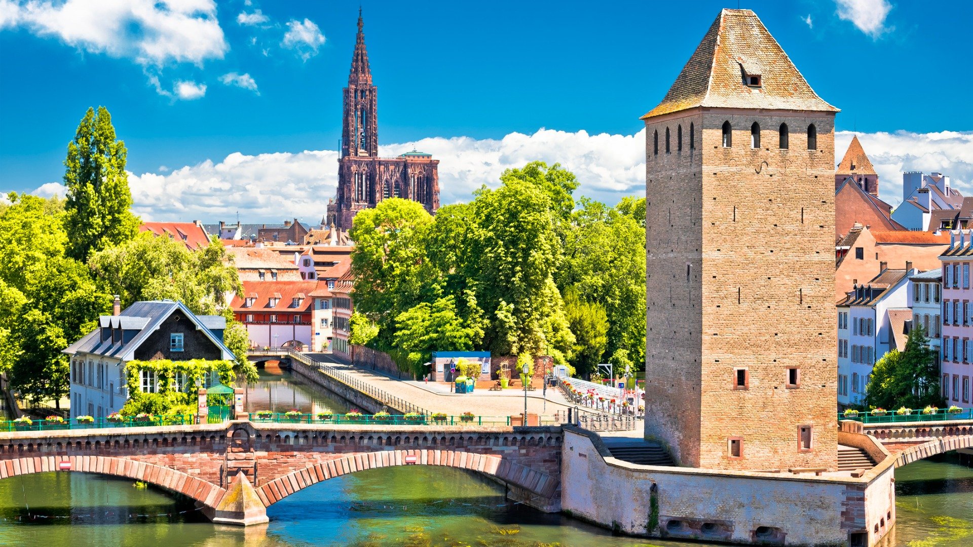 A tower in the foreground with a bridge, trees, and several buildings in the background. 